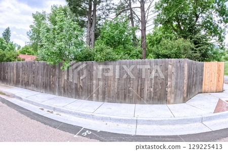 A tall wooden privacy fence forms a corner enclosure at the edge of a sidewalk. Trees and overgrown shrubs rise behind the fence, creating a secluded suburban feel. 129225413