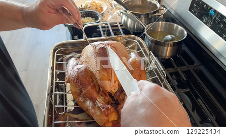 A person uses a baster to coat a golden brown roasted chicken inside a home oven. Roasting pans and spices are visible in the background. 129225434