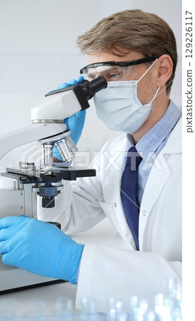 Masked male scientist analyzing sample under microscope, colleague researching nearby in sterile medical laboratory setting. Medicine, healthcare and science concept 129226117