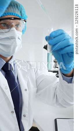 Scientist in protective gear analyzing a test tube of turquoise liquid with a pipette in a modern laboratory setting. Medicine, health care and science concept 129226118