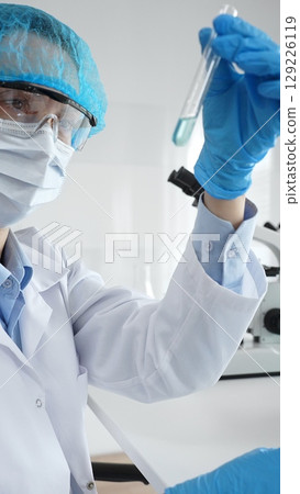 Close up of a scientist in ppe, holding a test tube with blue liquid while conducting research in a laboratory with a microscope. Medicine, health care and science concept Close up of a scientist in ppe, holding a test tube with blue liquid while conducting research in a laboratory with a microscope. Medicine, health care and science concept 129226119