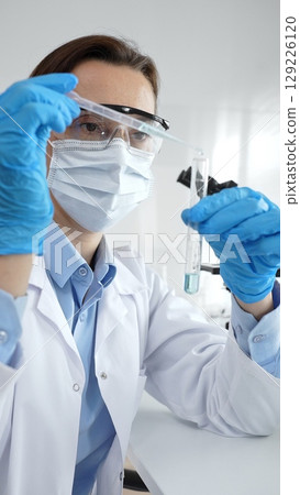 Close up of scientist wearing mask and gloves adding fluid to test tube using pipette in laboratory for medical research. Medicine, health care and science concept 129226120