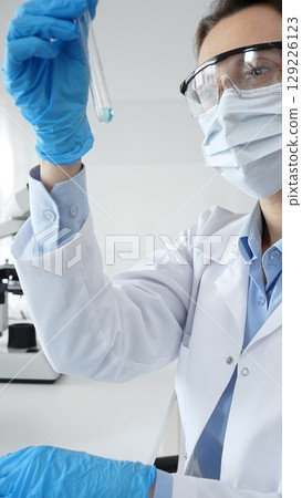 Female scientific researcher holding test tube, analyzing blue liquid while wearing protective gear during medical experiment in sterile laboratory setting. Medicine and science concept 129226123