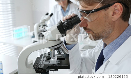 Medical scientist wearing gloves and protective glasses examining samples under a microscope in a bright laboratory filled with test tubes. Medicine, healthcare and science concept 129226133