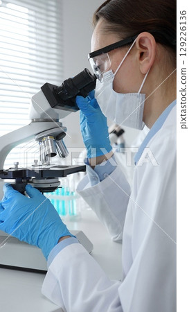 Female scientist wearing mask and blue gloves using microscope doing scientific research in modern laboratory with colleague working in background. Medicine, healthcare and science concept 129226136