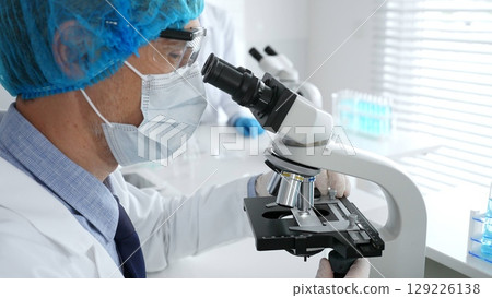 Senior male scientist wearing a protective mask, cap, and gloves, examining samples under a microscope in a modern laboratory. Medicine, healthcare and science concept 129226138