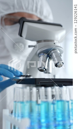 Scientific researcher or virologist wearing protective gear adjusting microscope, analyzing lab samples with precision in sterile research environment. Medicine and science concept 129226141