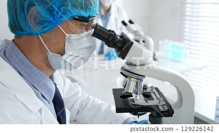 Senior male scientist wearing a protective mask, cap, and gloves, examining samples under a microscope in a modern laboratory. Medicine, healthcare and science concept 129226142
