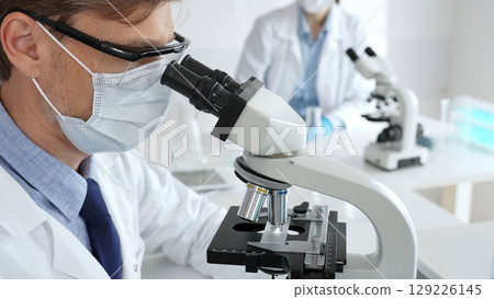Medical researcher analyzing sample under microscope, wearing protective gear in sterile laboratory setting, performing scientific research. Medicine, healthcare and science concept 129226145