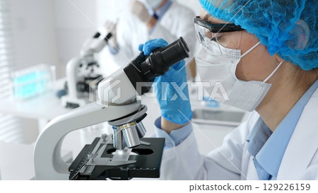 Female scientist wearing a mask and blue safety gear, using a microscope for research while a male colleague works in the background. Medicine, healthcare and science concept 129226159