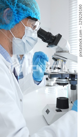 Senior male scientist wearing a protective mask, cap, and gloves, examining samples under a microscope in a modern laboratory. Medicine, healthcare and science concept 129226160