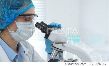 Female scientist analyzing sample through microscope, wearing protective lab gear during medical research investigation. Medicine, healthcare and science concept 129226167