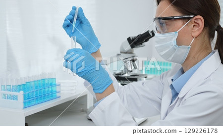 Female scientist wearing a face mask and blue gloves, carefully dropping liquid from a pipette into a test tube in a modern lab. Medicine, healthcare and science concept 129226178