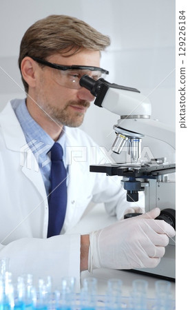 Researcher examining sample through microscope, wearing safety eyewear in sterile laboratory setting, surrounded by scientific glassware. Medicine, healthcare and science concept Researcher examining sample through microscope, wearing safety eyewear in sterile laboratory setting, surrounded by scientific glassware. Medicine, healthcare and science concept 129226184
