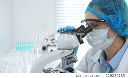 Female scientist analyzing sample through microscope, wearing protective lab gear during medical research investigation. Medicine, healthcare and science concept 129226185