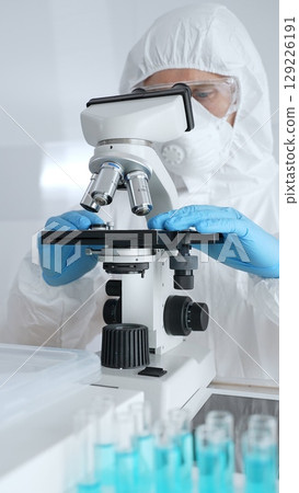 Scientific researcher wearing protective gear adjusting microscope, analyzing lab samples with precision in sterile research environment. Medicine, healthcare and science concept 129226191