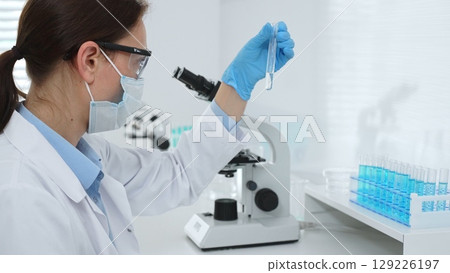 Female researcher wearing protective mask and blue gloves is examining liquid in test tube, sitting near microscopes and lab equipment in laboratory. Medicine, healthcare and science concept 129226197