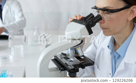 Female scientist examining sample under microscope, conducting research in laboratory with colleague working near test tube rack. Concept of medicine, healthcare and science Female scientist examining sample under microscope, conducting research in laboratory with colleague working near test tube rack. Concept of medicine, healthcare and science 129226206