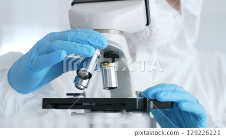 Close up of scientific researcher or virologist wearing protective gear is adjusting microscope, analyzing lab samples with precision in sterile research environment. Medicine and science concept Close up of scientific researcher or virologist wearing protective gear is adjusting microscope, analyzing lab samples with precision in sterile research environment. Medicine and science concept 129226221