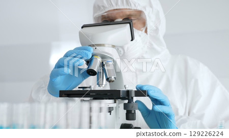 Male scientific researcher or virologist wearing protective gear adjusting microscope, analyzing lab samples with precision in sterile research environment. Medicine concept Male scientific researcher or virologist wearing protective gear adjusting microscope, analyzing lab samples with precision in sterile research environment. Medicine concept 129226224