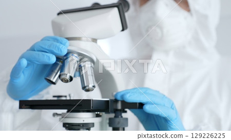 Close up of scientific researcher or virologist wearing protective gear is adjusting microscope, analyzing lab samples with precision in sterile research environment. Medicine and science concept Close up of scientific researcher or virologist wearing protective gear is adjusting microscope, analyzing lab samples with precision in sterile research environment. Medicine and science concept 129226225