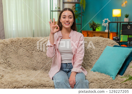 Young woman making confident okay hand gesture smiling to camera showing approval and trust on sofa Young woman making confident okay hand gesture smiling to camera showing approval and trust on sofa 129227205