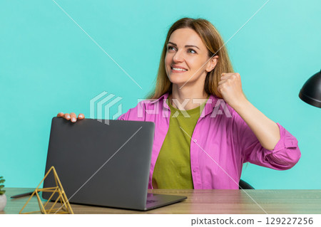 Woman sitting at table celebrating victory or success after receiving great news on laptop screen Woman sitting at table celebrating victory or success after receiving great news on laptop screen 129227256