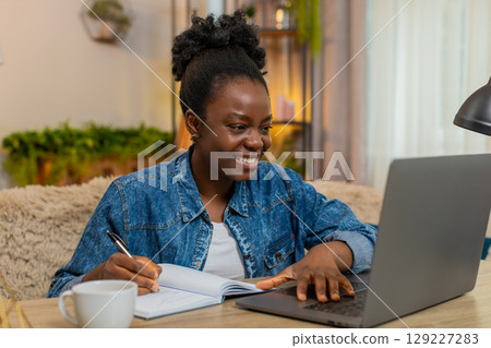 African American young woman studies online with laptop and notebook takes write notes at home table 129227283