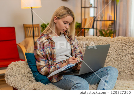Adult woman writing financial notes in notebook while looking at laptop screen at home with focus Adult woman writing financial notes in notebook while looking at laptop screen at home with focus 129227290