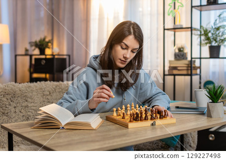 Young woman playing chess alone arranging pieces at table on sofa with book lying nearby at home 129227498