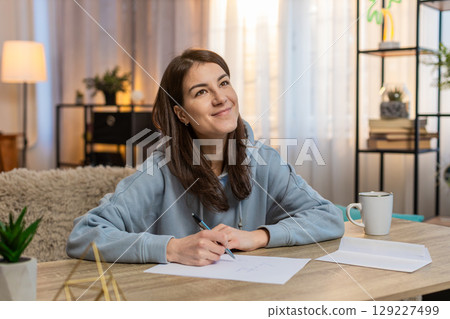 Happy woman sitting on sofa at table deeply focused on writing important letter by hand at home Happy woman sitting on sofa at table deeply focused on writing important letter by hand at home 129227499