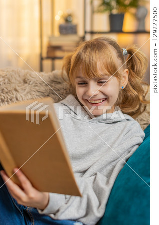 Young small teenager girl reading favorite storybook at home sofa quiet smiles, tiny serious sighs 129227500