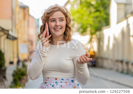 Happy young woman having remote conversation talking on smartphone good news gossip in city street 129227508