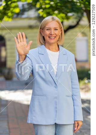 Happy mature businesswoman looking at camera waving hello greeting gesture friendly on city street 129227509