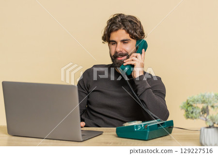 Happy businessman sitting at table talking on wired telephone and using laptop on beige background 129227516