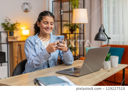 Smiling young woman freelancer sitting at table using smartphone texting in living room at home 129227528