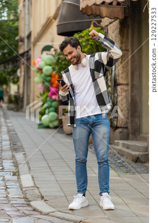 Hispanic young man using smartphone celebrating win good message news clenching fist on city street Hispanic young man using smartphone celebrating win good message news clenching fist on city street 129227533