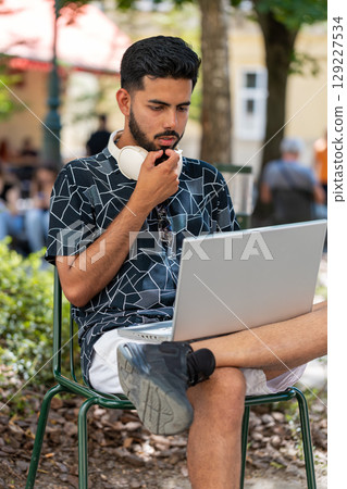 Indian man in headphones working online distant job with laptop browsing website chatting outdoors Indian man in headphones working online distant job with laptop browsing website chatting outdoors 129227534