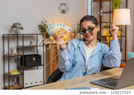 Young woman freelancer putting on party glasses holding fan of cash sitting at home office table Young woman freelancer putting on party glasses holding fan of cash sitting at home office table 129227535