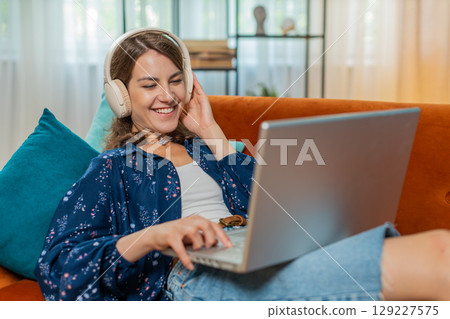 Happy woman in wireless headphones lying on home couch listening music working on laptop relaxing 129227575