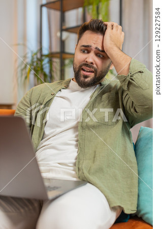 Upset young Caucasian young man sitting on sofa at home using laptop having anxiety and stress Upset young Caucasian young man sitting on sofa at home using laptop having anxiety and stress 129227584