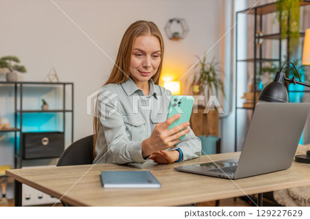 Happy female freelancer sitting at desk using smartphone texting smiling in living room at home 129227629