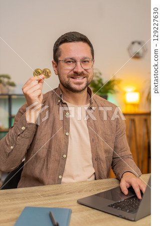 Portrait of happy young male freelancer trader showing golden bitcoins sitting at home office table 129227630