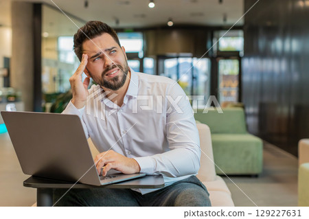 Tired ill young businessman using laptop suffering from headache problem tension in hotel lobby 129227631