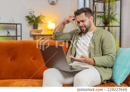 Upset young Caucasian man freelancer sitting on sofa at home using laptop having anxiety and stress 129227649