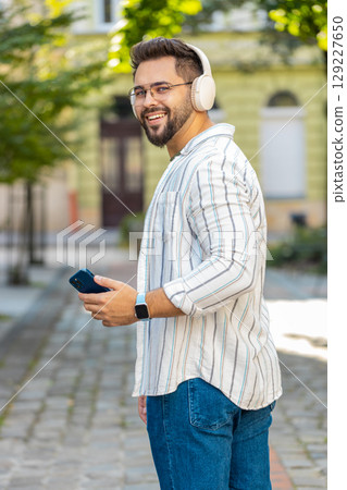 Happy young man in glasses listening music via wireless headphones while walking on city street 129227650