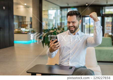 Happy young businessman using smartphone shocked by sudden victory lottery win in office lobby 129227658