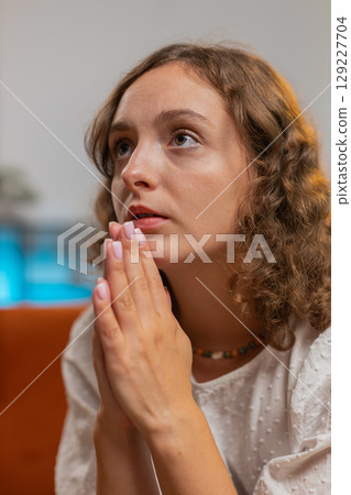 Sad woman sitting on sofa at home in shock by awful news, folding palms together pray ask for help 129227704