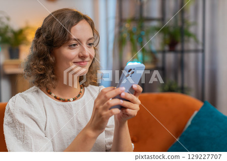 Happy young woman sitting on sofa using smartphone texting smiling at home living room apartment 129227707