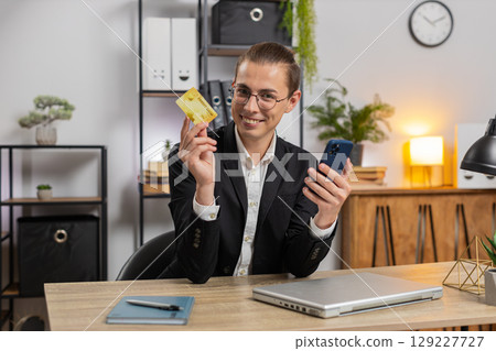 Happy young businessman using bank credit card and smartphone for online shopping at home office Happy young businessman using bank credit card and smartphone for online shopping at home office 129227727